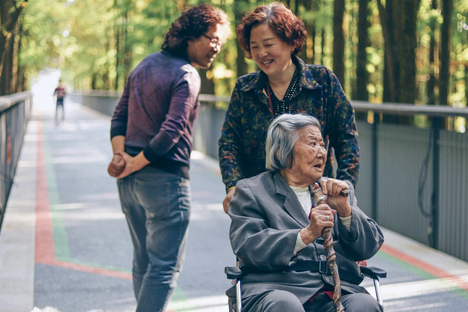 An older woman using a wheelchair outdoors with two supportive companions nearby